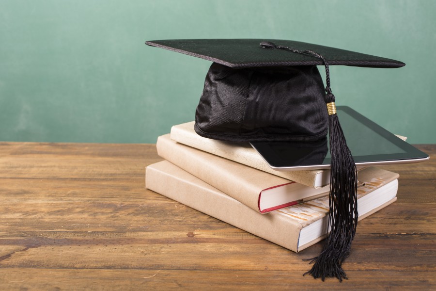 Spring graduation time. A black cap/motarboard with black tassel lies on top of a stack of textbooks and digital tablet. All objects lie on a rustic wooden desk with green chalkboard in background. Copyspace to side. No people. Education themes.