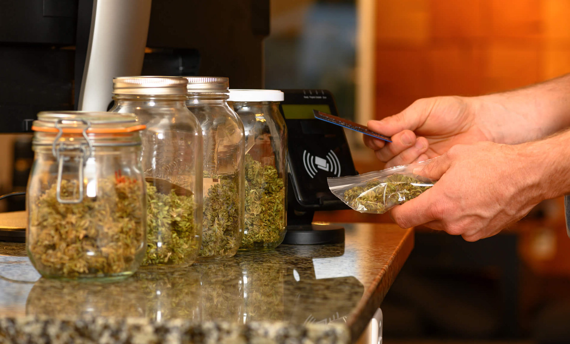 Jars of cannabis flower at a dispensary with a customer's hands using a credit card to purchase