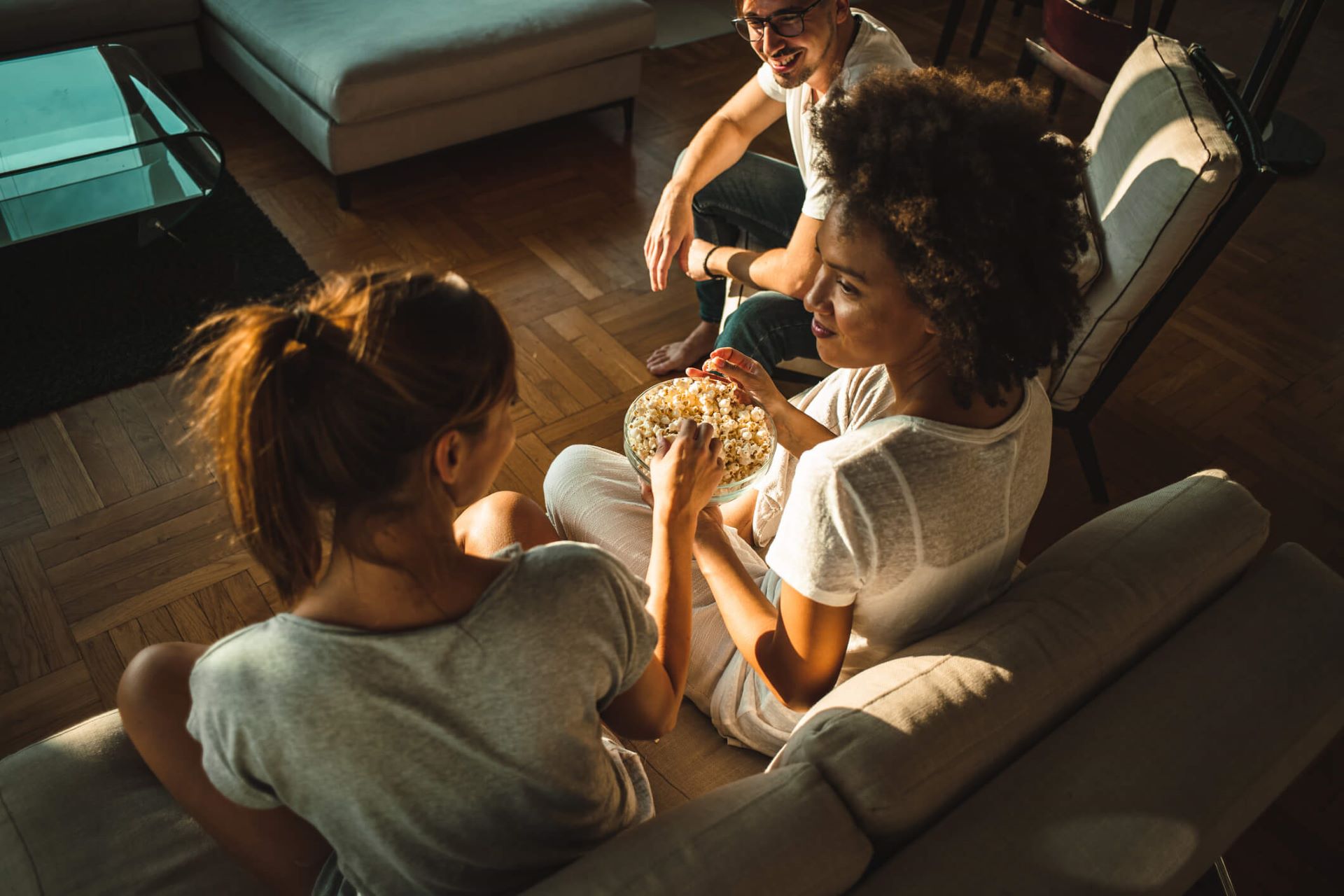 An image showing three friends watching TV in the living room while eating popcorn.