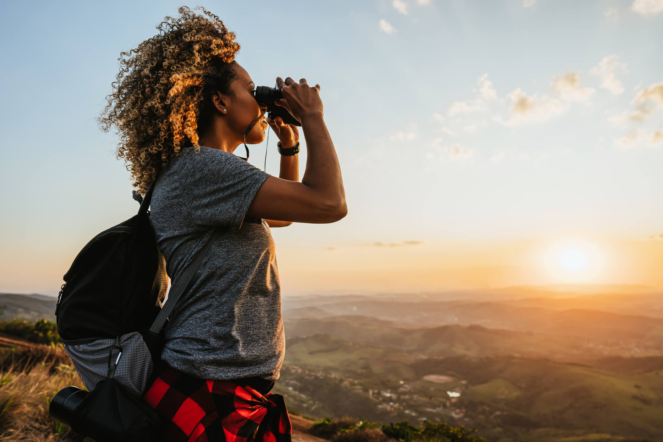 A woman on top of a ridge or mountain looking through binoculars with the sunrise in the background.