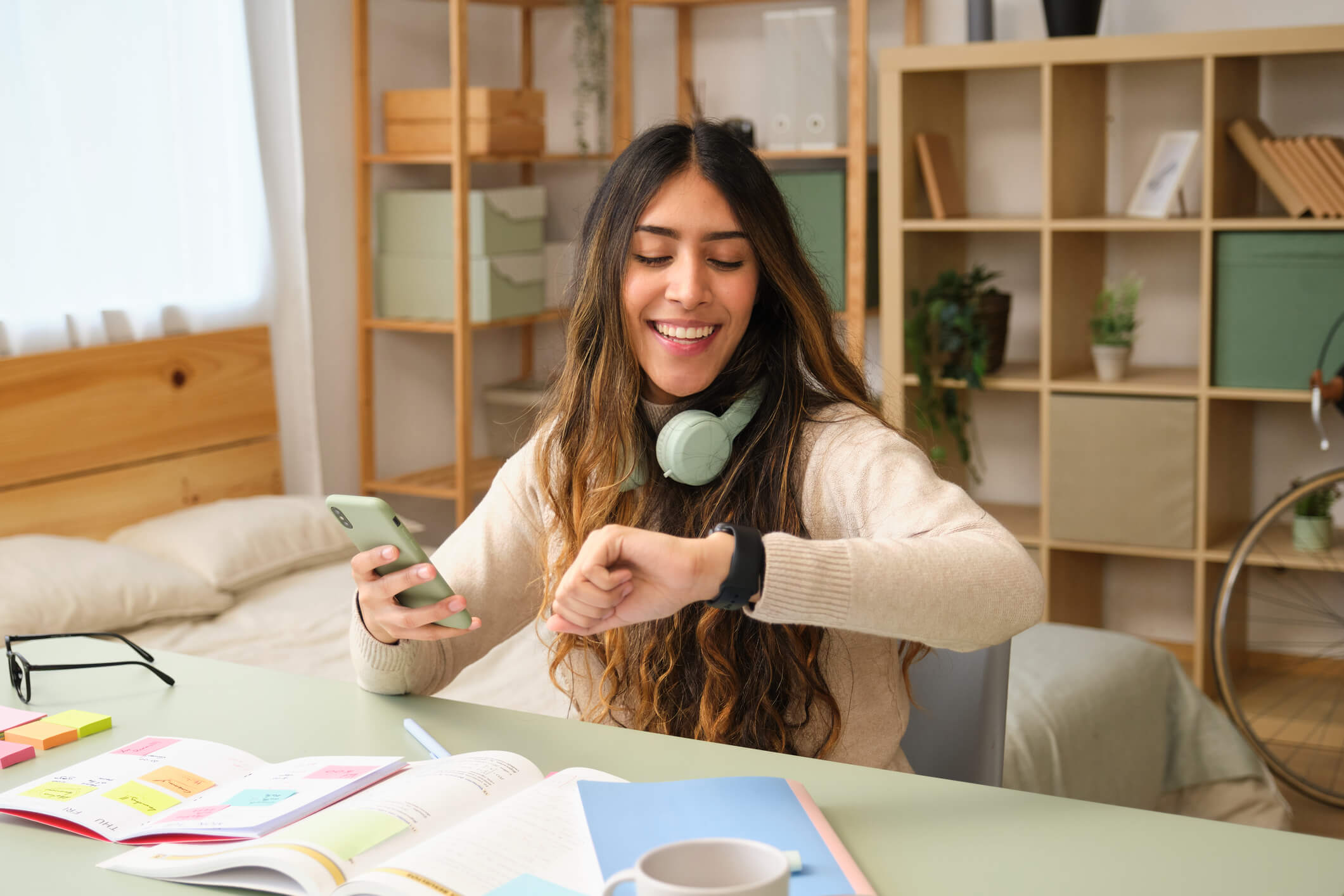 Distracted woman with headphones on sits at desk while looking at watch and holding phone