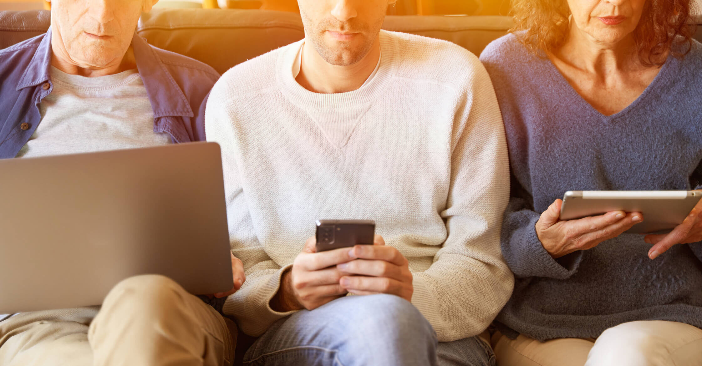 Three people sitting on a couch watching different devices: A laptop, a phone, and a tablet.